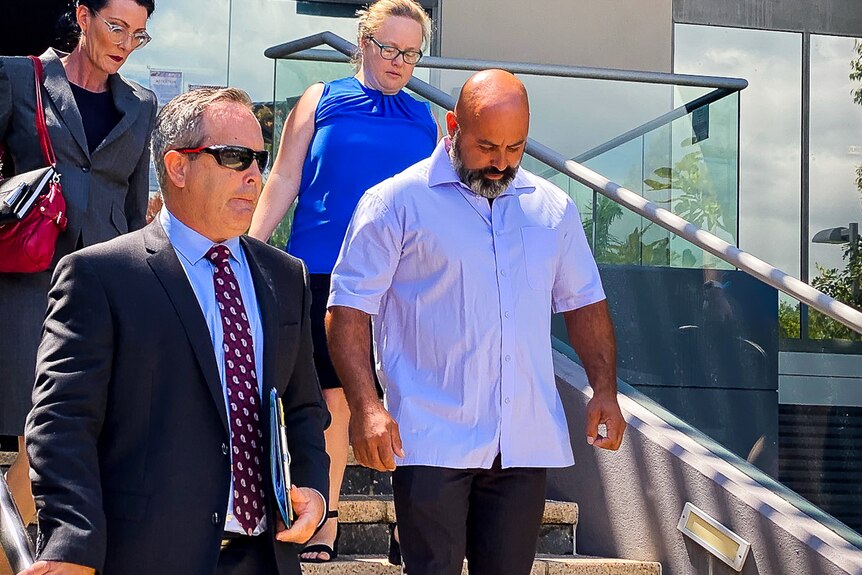 Man walking down stairs wearing purple shirt and black slacks, surrounded by three members of his legal team