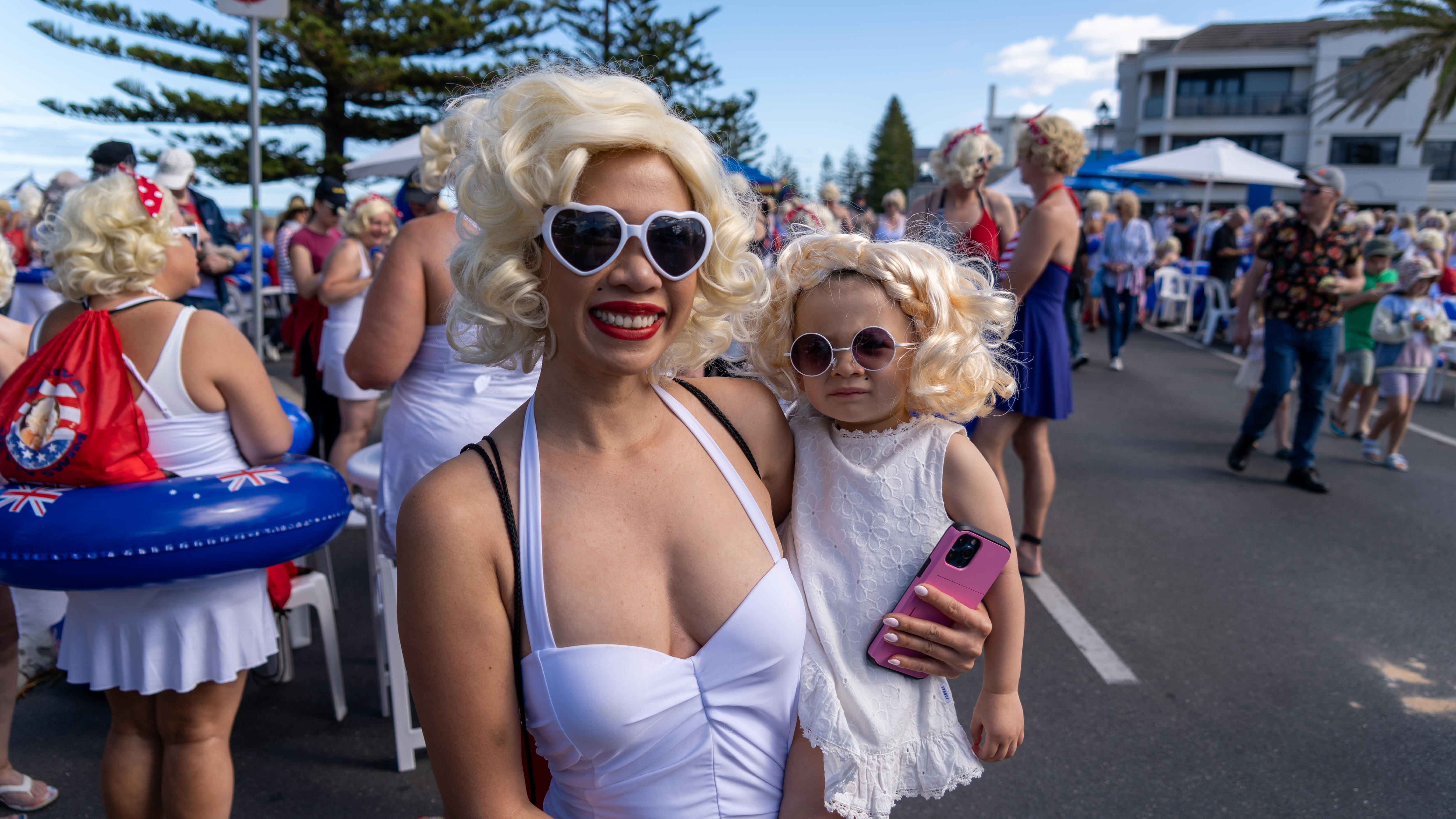 A woman holds a small child on her hip, both are dressed in white, with sunglasses and curly blonde wigs