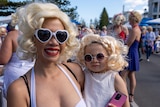 A woman holds a small child on her hip, both are dressed in white, with sunglasses and curly blonde wigs