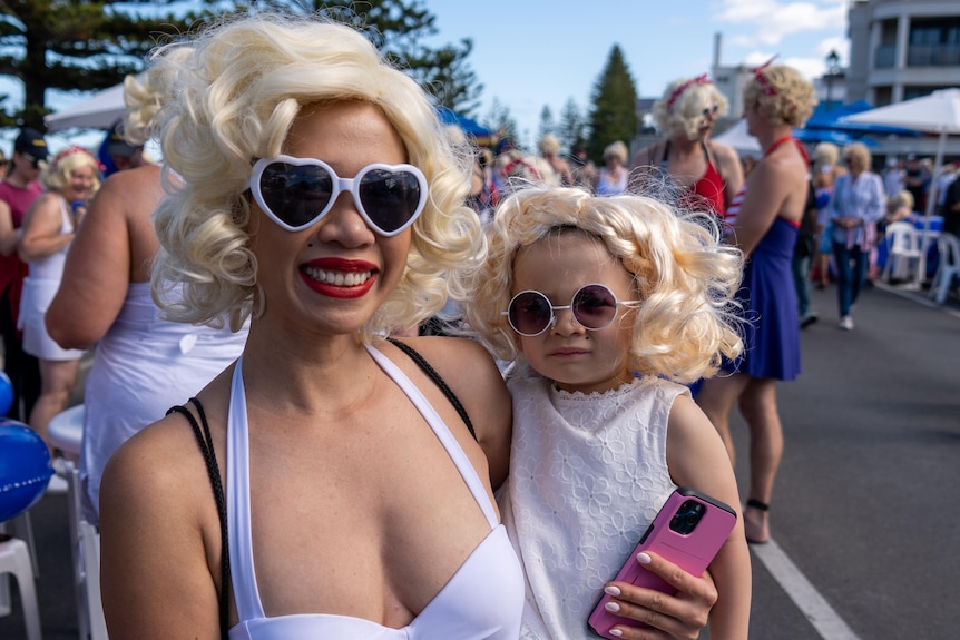 A woman holds a small child on her hip, both are dressed in white, with sunglasses and curly blonde wigs