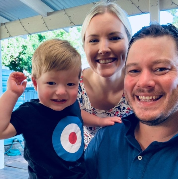 A blonde haired toddler being held by a man in a blue shirt with a blonde woman standing behind them