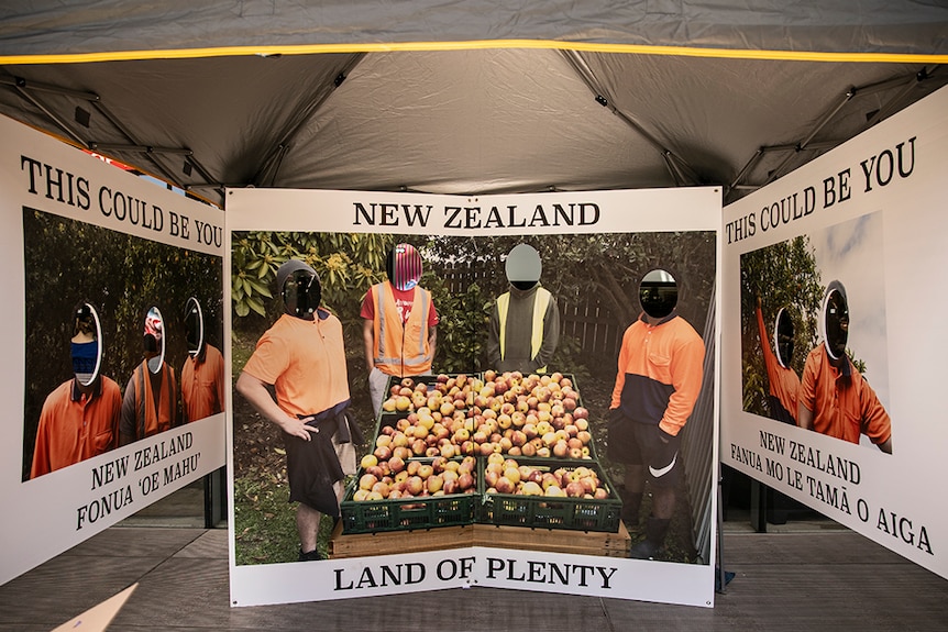 Three photo boards with text and pictures of labourers with faces cut out hang under marquee tent.