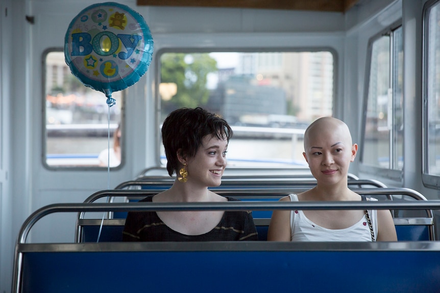 Two women sit together on an empty bus, the woman on the left smiles while holding a helium balloon the reads 'boy'.