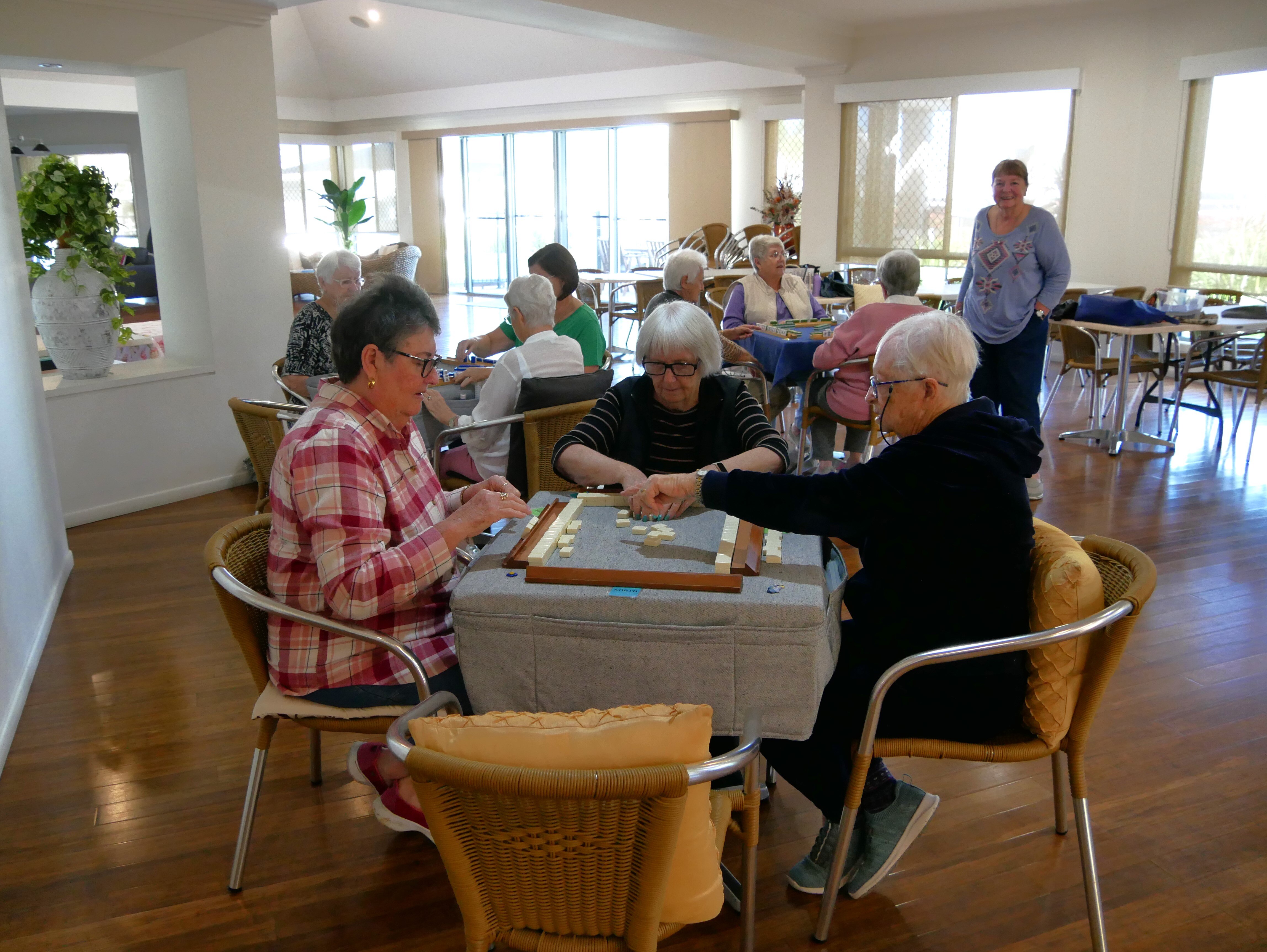 Groups of older women sitting at tables and chairs playing mahjong at a clubhouse. 