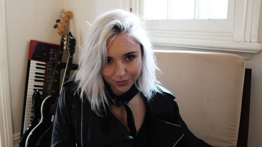 Noelle Faulkner sitting in a chair, she has platinum-blonde hair and is wearing a leather jacket.