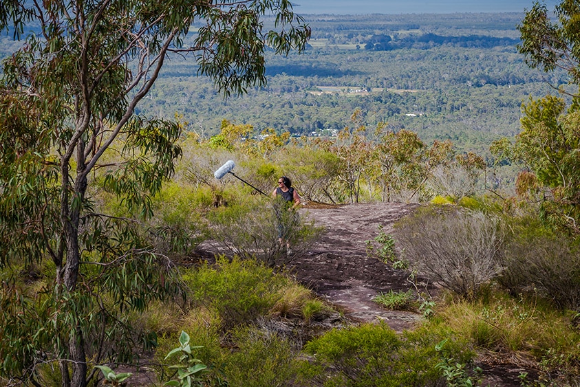 Dr Leah Barclay with a boom recorder on an escarpment in Australian bushland overlooking a valley.