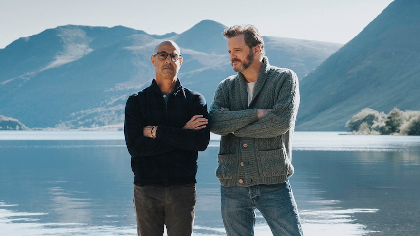 Two middle-aged men in woollen jumpers and jeans stand before a lake in the mountains with arms crossed.