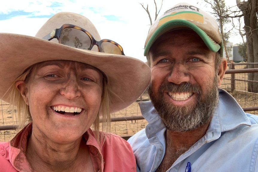 A man and woman smile in cattle yards, covered in dust. 