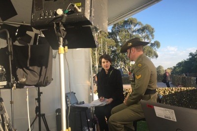 Trioli doing interview with man in soldier's uniform and memorial in background.