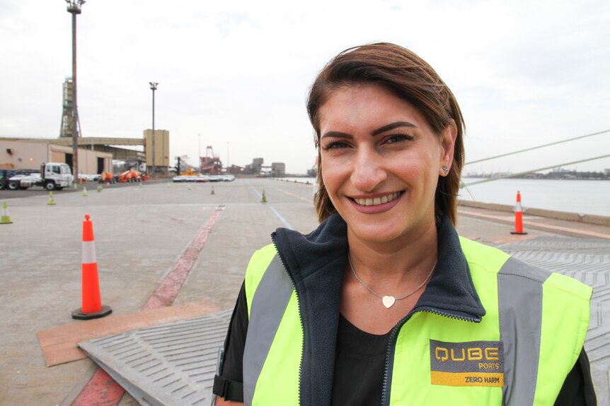 A brown-haired woman in a high-vis vest smiles directly at the camera on a dockside