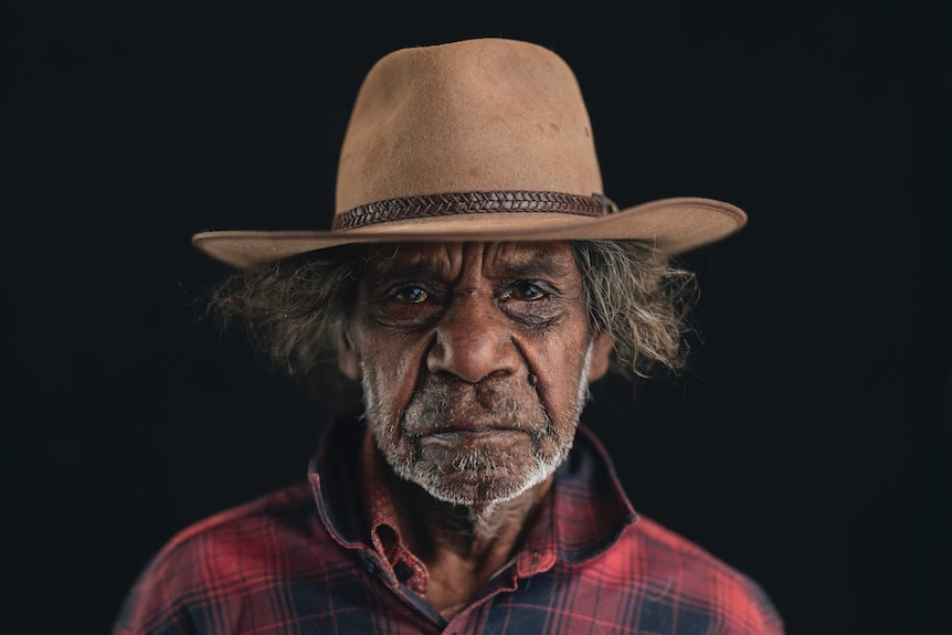An Aboriginal man wearing a tan hat and checked red shirt looks toward the camera against a black background.