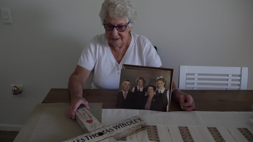 A woman in her 80s sits at a kitchen table with the cross in one hand and a photo of her father stands next to it.