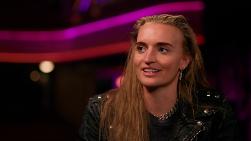 G Flip on the right hand side of the screen with their hair out wearing a black leather jacket, the background is out of focus