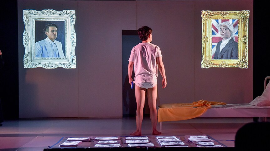 A man in white t-shirt and underwear stands with back to audience and looks to two ornate framed pictures projected onto stage.