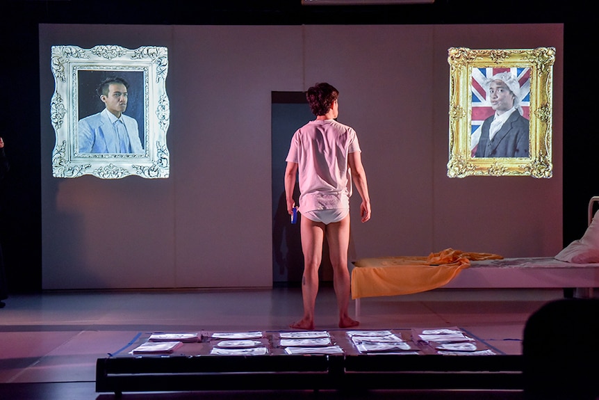A man in white t-shirt and underwear stands with back to audience and looks to two ornate framed pictures projected onto stage.