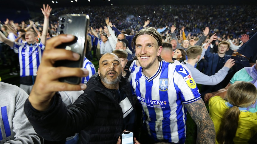 An excited football fan takes a selfie next to a smiling footballer from his team after a big win.