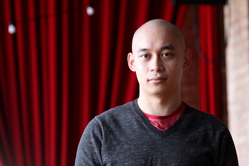 A man looks at the camera, red curtain in background.