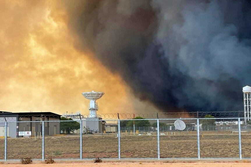 Large plumes of black smoke erupt behind a fenced off military base