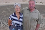 A couple stand in front of a dry and empty dam on a farm.
