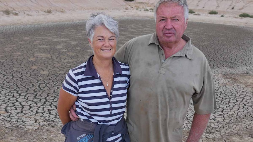 A couple stand in front of a dry and empty dam on a farm.