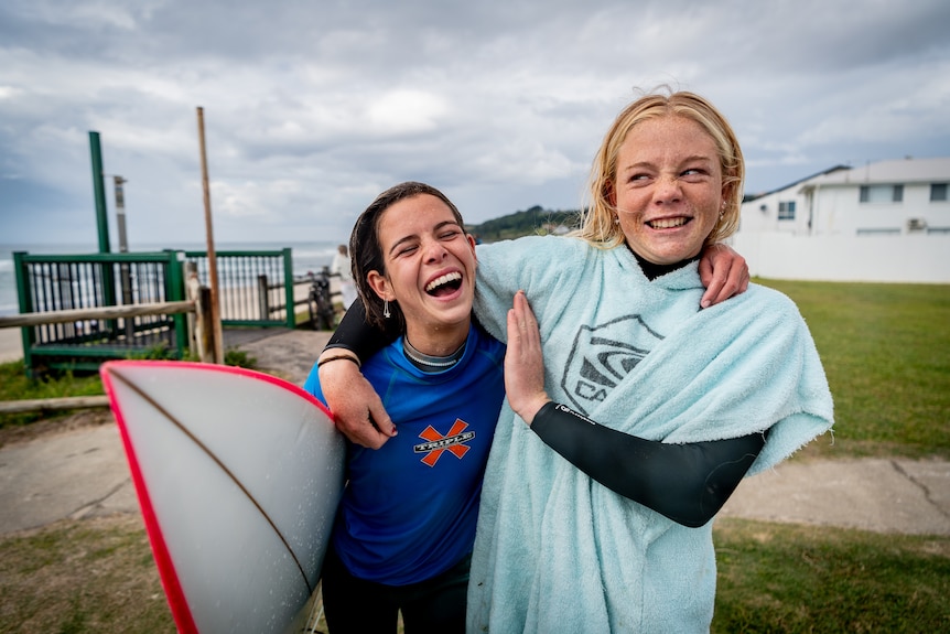 Two young girls arm in arm pulling faces and laughing. One has a surfboard under their arm.