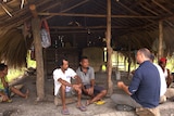 Mark Willacy sits and speaks with two Timorese men in a thatched hut.