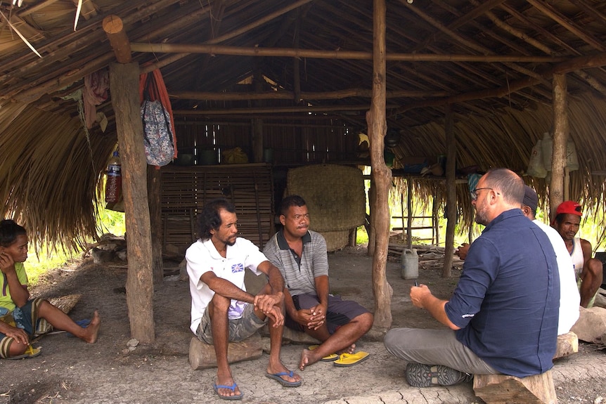 Mark Willacy sits and speaks with two Timorese men in a thatched hut.