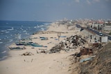 A wide photo of a long beach, with small boats, rubble and sheets of metal on the sand. Cars drive on a road nearby