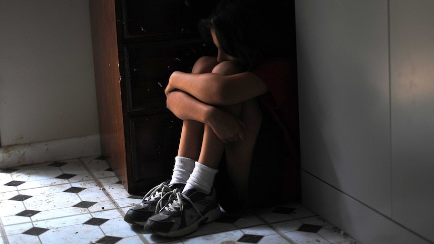 A girl sits on the floor hunched in a corner next to a cupboard, peering over her knees.