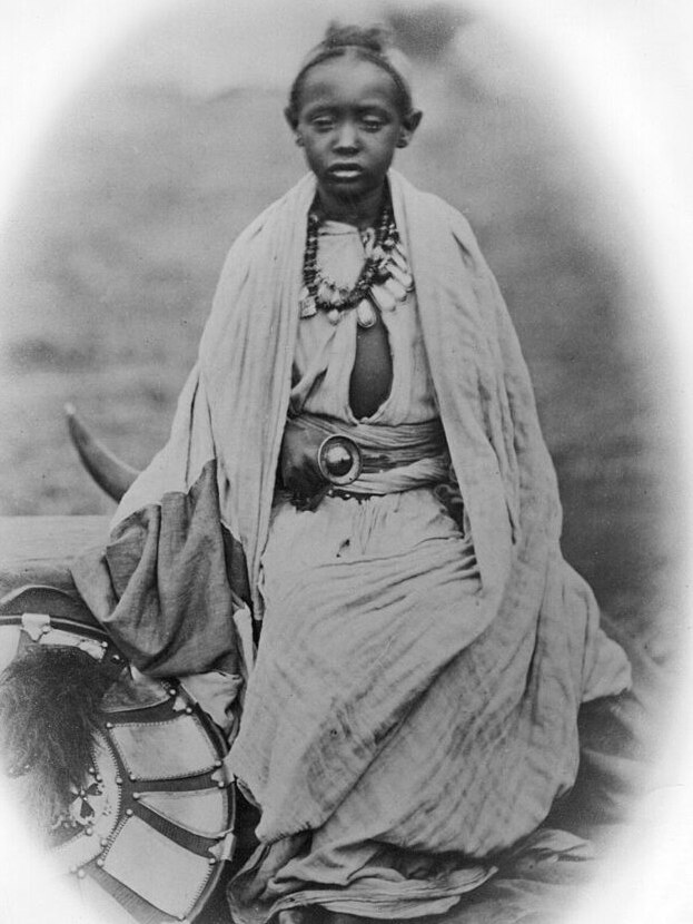 Photograph of a young boy with Ehtiopian clothing dating back to the mid 180s. He has a necklace on and a shield nearby. 