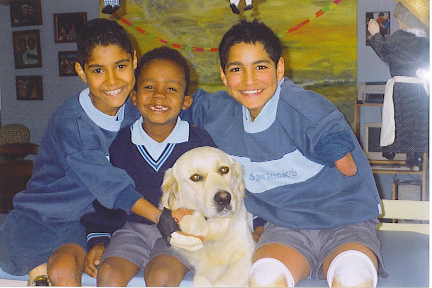 Three young boys in school uniforms smile at the camera next to a golden retriever dog.