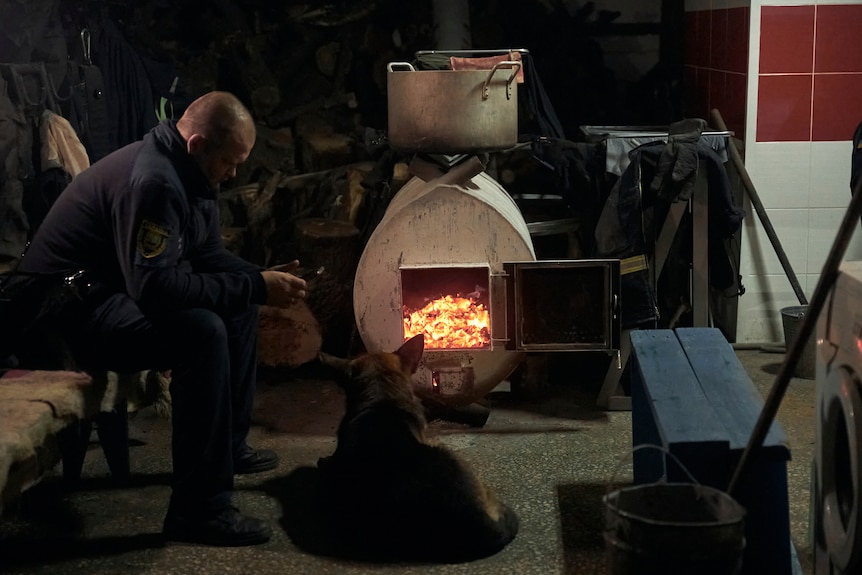 An emergency worker and his dog warm up in front of a wood-burning oven in a shelter.