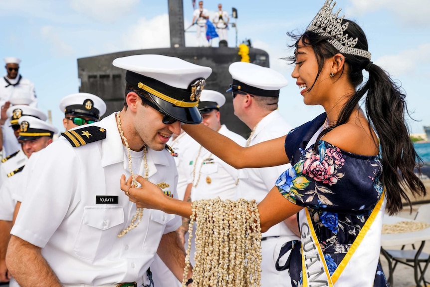A woman welcomes a US submariner by placing a shell necklace on him. She is a Miss Guam winner and is wearing a tiara.