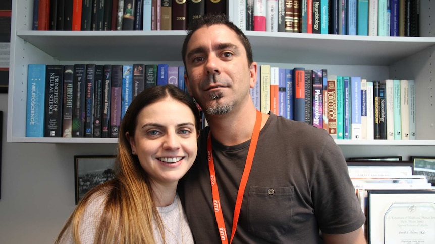 A young couple pose for the camera in front of bookshelves.