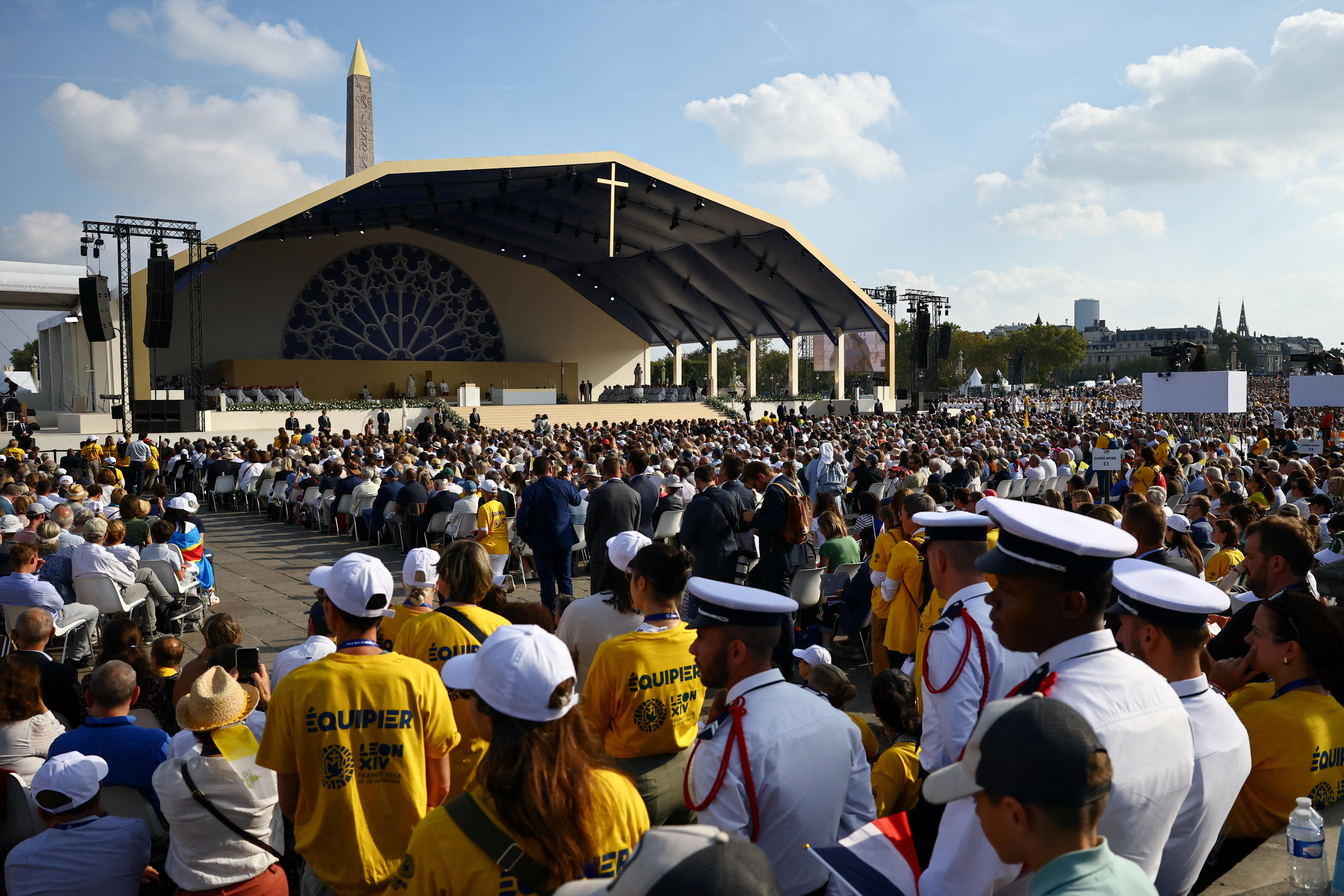 A large crowd before a stage with a cross near the roof.