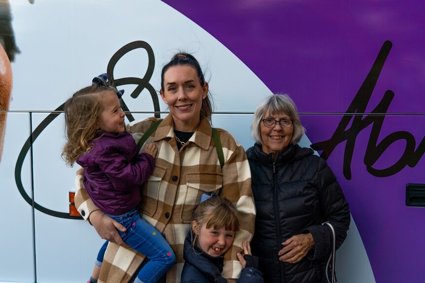 A woman in a brown-and-white checked jacket stands with her mum and two daughters in front of a purple Stacey Abrams bus