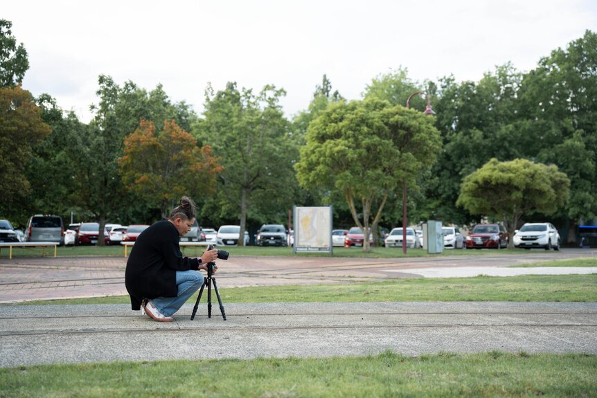 A woman squats down behind a camera on a tripod