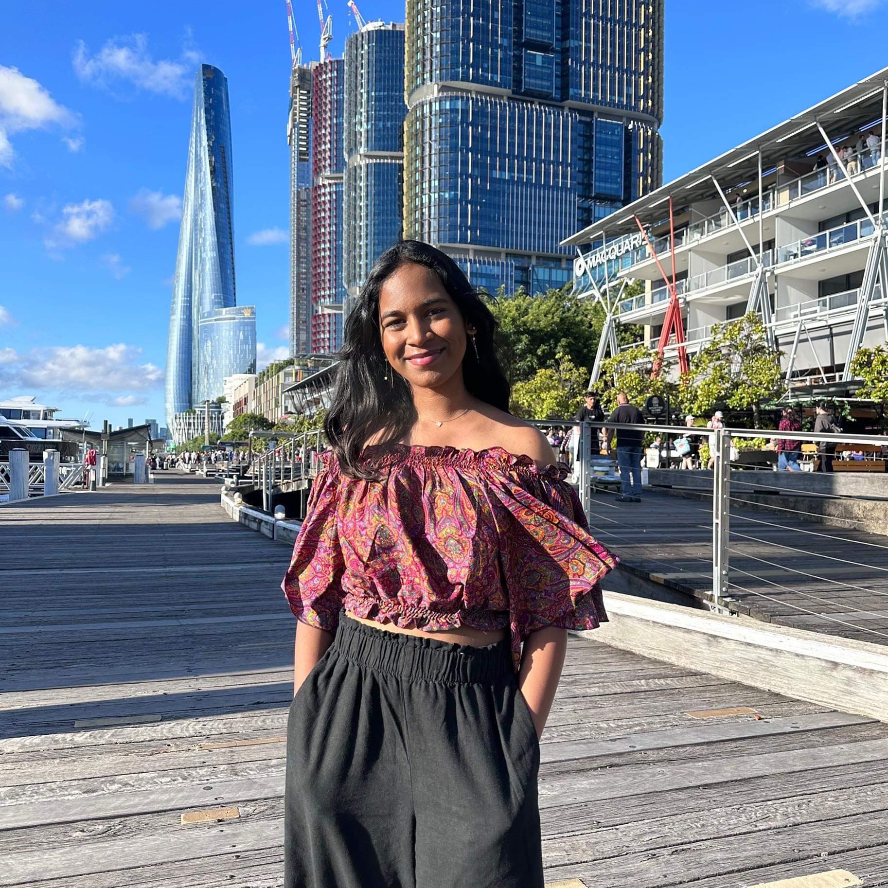 A photo of Satara standing in front of a few buildings. She's wearing an off the shoulder dark red top and black pants.
