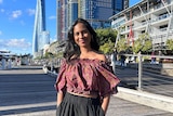 A photo of Satara standing in front of a few buildings. She's wearing an off the shoulder dark red top and black pants.