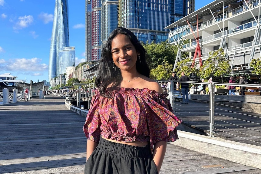 A photo of Satara standing in front of a few buildings. She's wearing an off the shoulder dark red top and black pants.