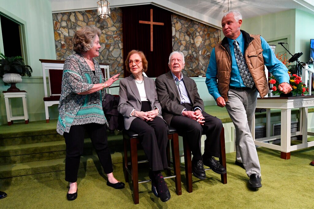 Former president Jimmy Carter and his wife sit after getting their photo taken at Sunday school, with a wooden cross behind them