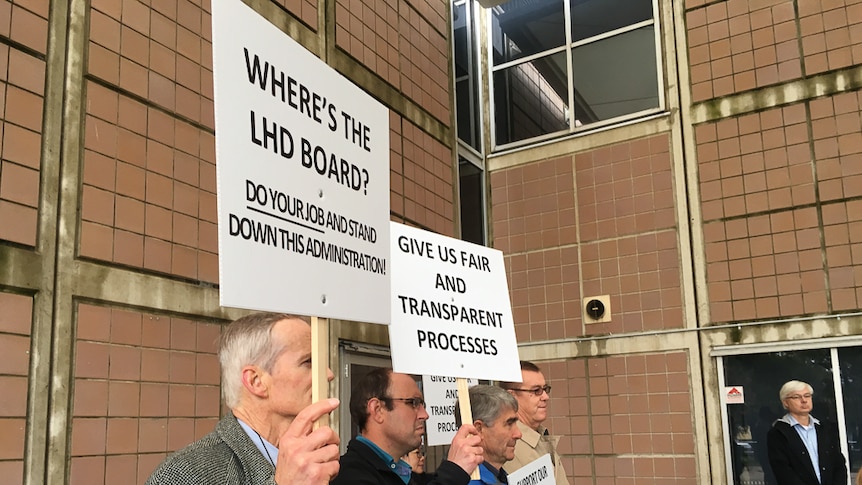 Side view of men standing holding signs saying Where's the LHD Board, give us fair and transparent process