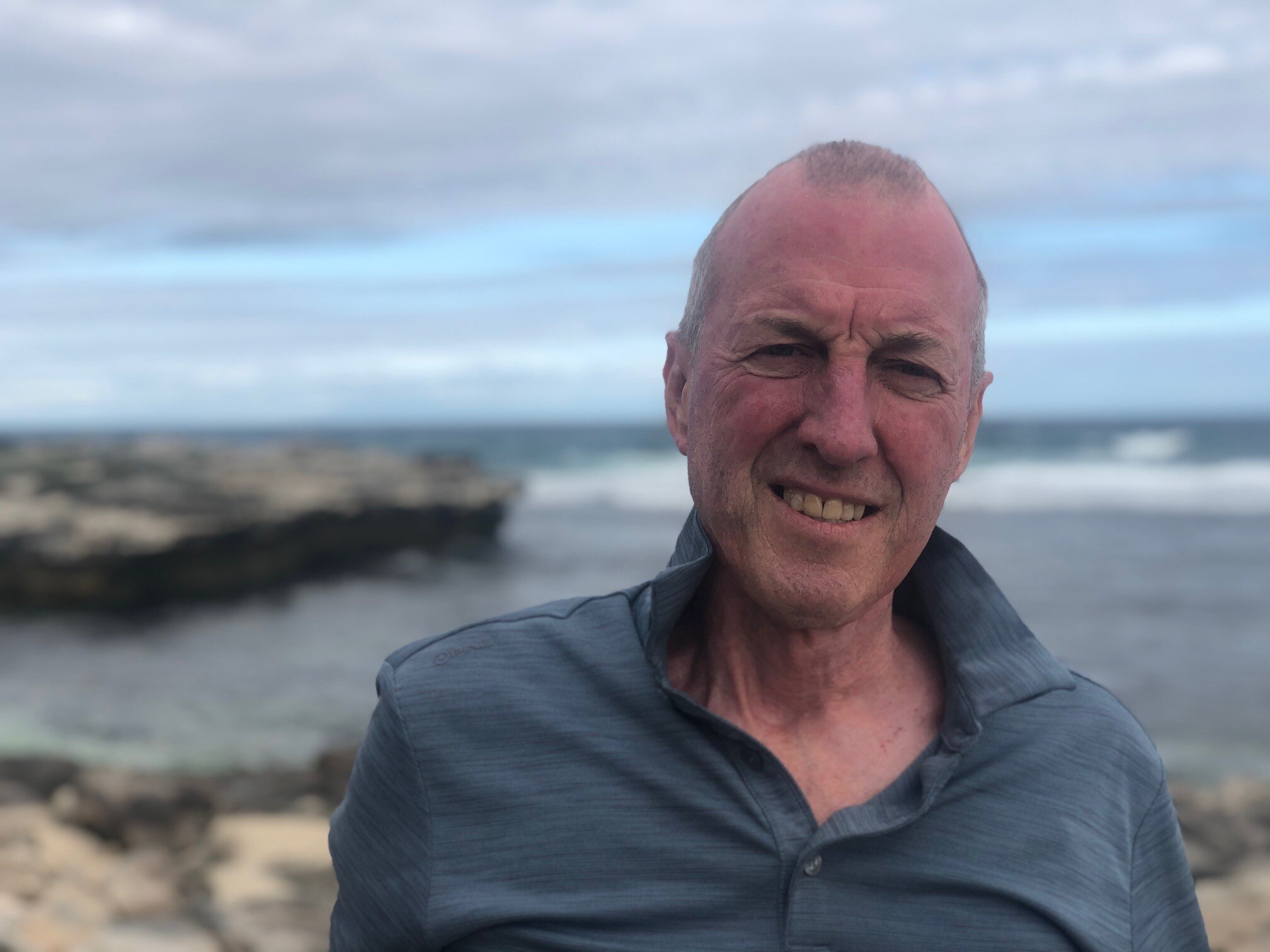 A close up of Mark wearing a dark grey/blue long sleeved shirt, with a coastal cliff behind him and ocean in the back.