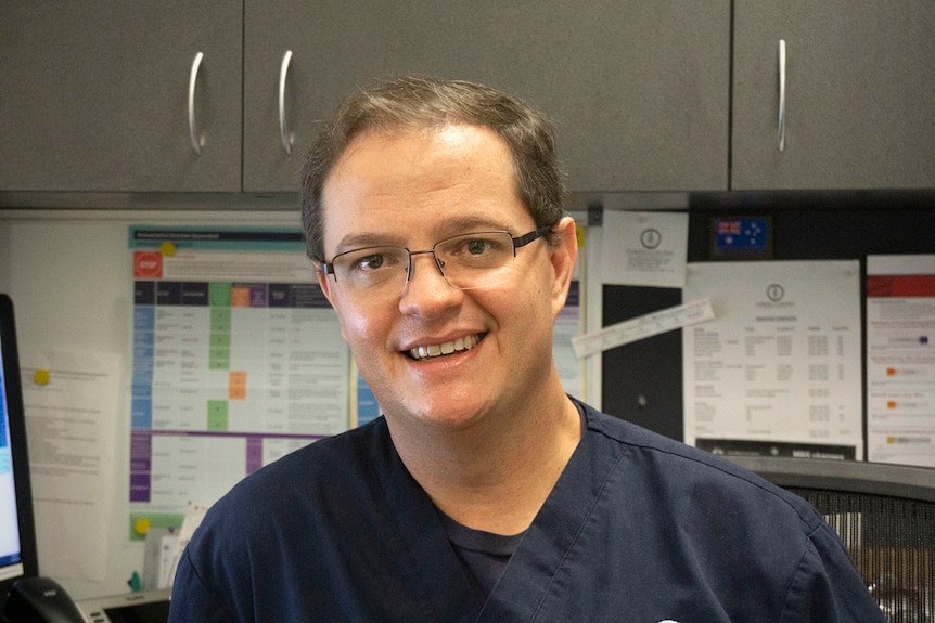 Un médico varón sentado en una silla y sonriendo a la cámara.  Lleva gafas y un uniforme azul oscuro.