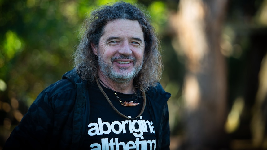 A man with long wavy hair and beard smiling to camera, t shirt says aboriginal all the time.