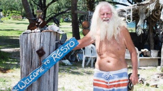 A man with long white hair and beard, wearing boardshorts, stands next to wooden painted sign saying 'Welcome to Resto'.