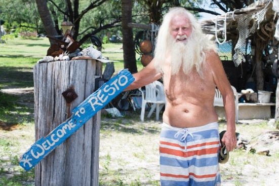 A man with long white hair and beard, wearing boardshorts, stands next to wooden painted sign saying 'Welcome to Resto'.