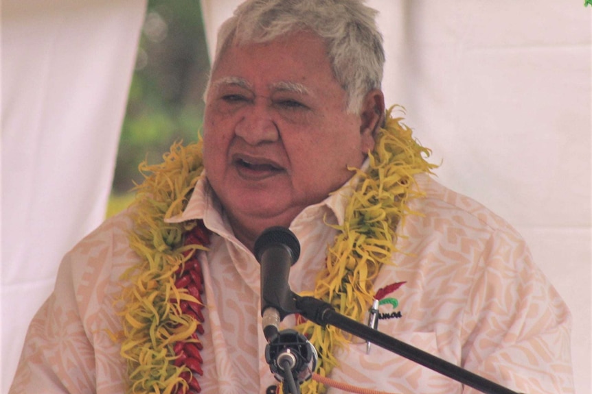 A plump, older man with silver hair wears a floral wreath around his neck as he speaks to a crowd from behind a lecturn.