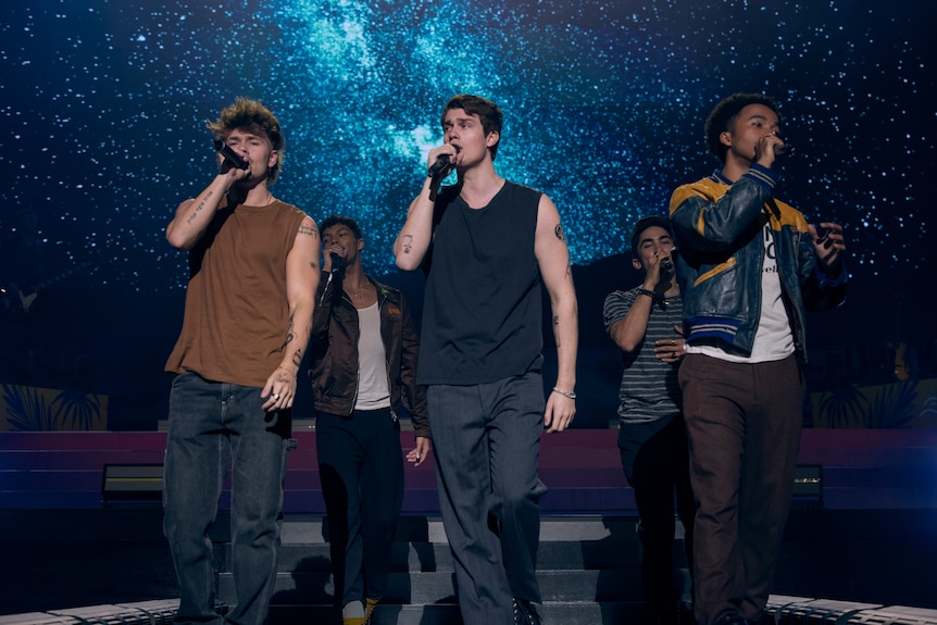 A film still of five young men, including Nicholas Galitzine, in a boy band performing on stage, mics to their mouths.