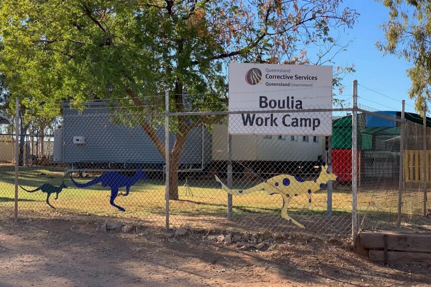 A group of dongas behind a fence with barbed while on the top, with a sign reading 'Boulia Work Camp'.